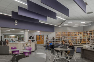 Common area in the new library at Odyssey High School, with circle tables, students studying and working together, a reception desk, and bookshelves on an elevated area in the back.