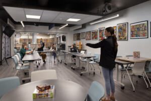 The improved makerspace at Odyssey High School with walls decorated with colorful student artwork, someone standing at the head of the room calling on a student, sitting with others in a loop of dynamic tables.