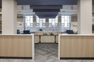 A large, semicirclular reception desk with different monitors and chairs behind the desk, filing cabinets, and columned tall windows across the room at Odyssey High School's renovated library.