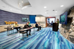 R21_14773_01 A classroom with blue carpeting reminiscent of ocean water, people sitting at a few tables and someone at a podium next to a monitor on a rocky wall at the head of the room.