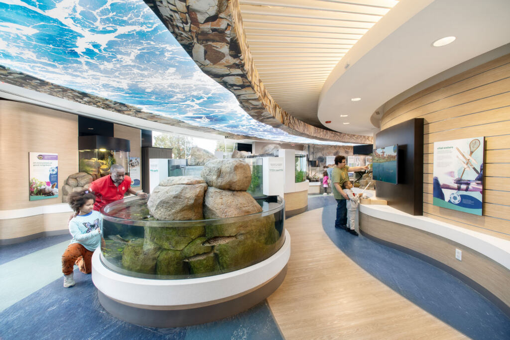 A wide view of people exploring the aquarium exhibits, including fish tanks, wall displays, monitors, and lake-themed floor and ceiling designs.
