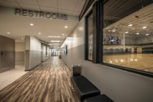A hallway with windows looking into a gymnasium, as well as overhead navigational signs reading "RESTROOMS" and "CONCESSIONS".
