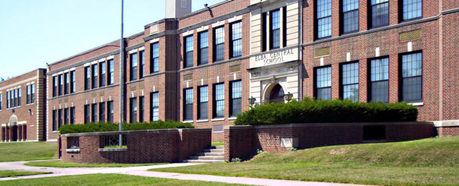 Front view of Elba Central School District’s historic brick school building with American flag flying on the lawn under a clear blue sky.