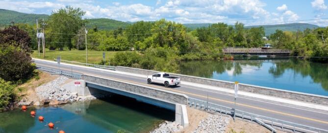 A newly constructed bridge carrying a pickup truck over a calm river, with an older, deteriorating bridge visible in the background—illustrating the replacement of aging infrastructure to enhance transportation safety and resilience.