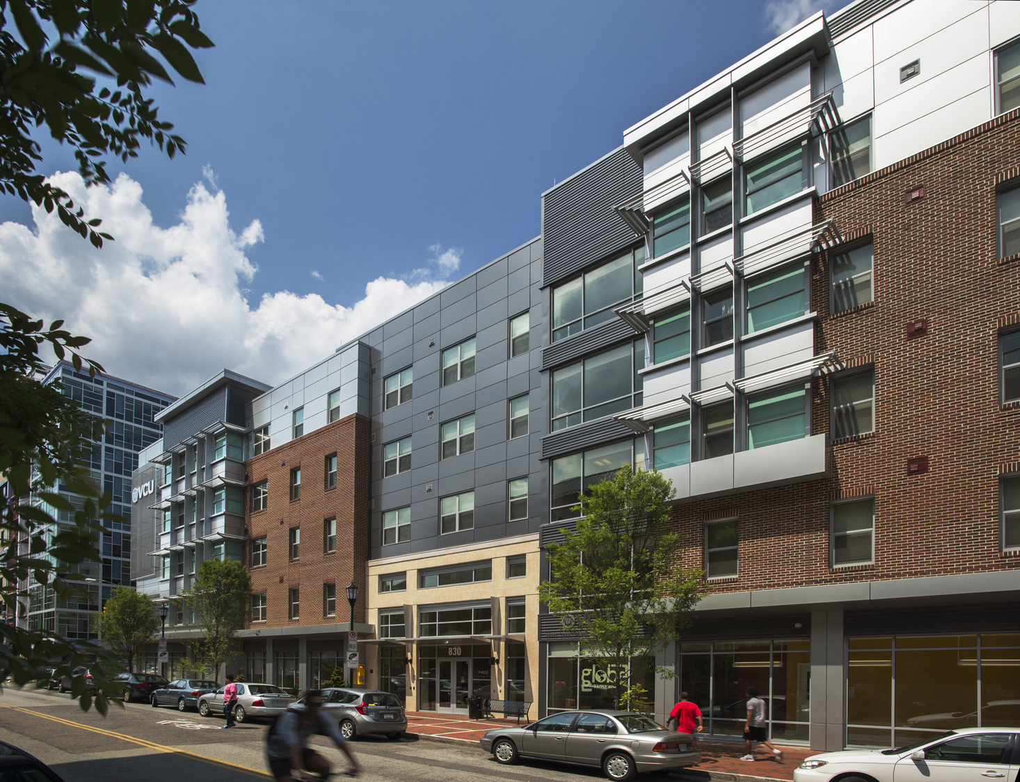 Exterior VCU student housing. Modern urban street with a mix of brick and glass buildings under a bright blue sky. Cars parked and pedestrians walking on the sidewalk convey a lively city atmosphere.