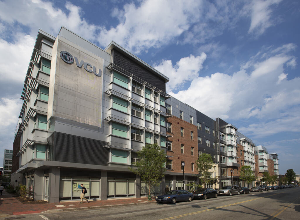 Exterior of Virginia Commonwealth University Student Housing. Modern multi-story building with a mix of glass and brick facades, VCU logo visible. A person walks on the sidewalk under a partly cloudy blue sky.