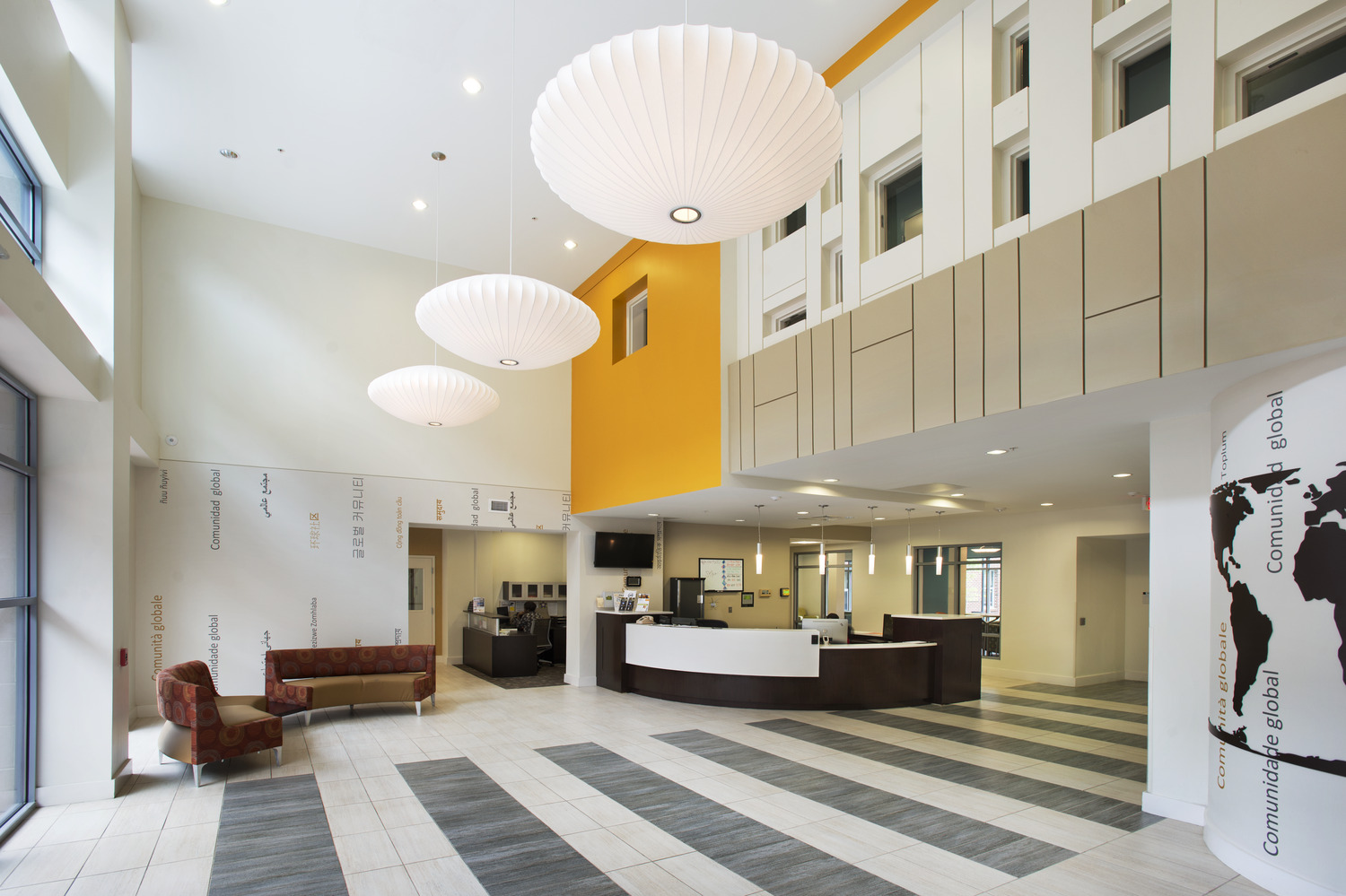 Spacious modern lobby at Virginia Commonwealth University with high ceiling, large round pendant lights, white and orange walls, and a central reception desk. Brown seating area to the left.