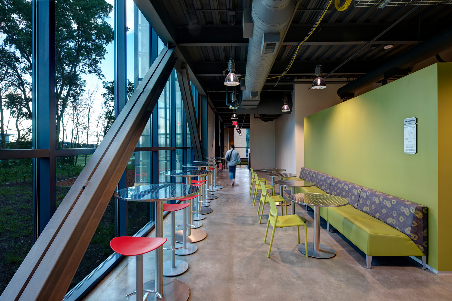 A hallway with high tables and stools flanking a glass wall, and tables and booths against a lime green wall on the other side.
