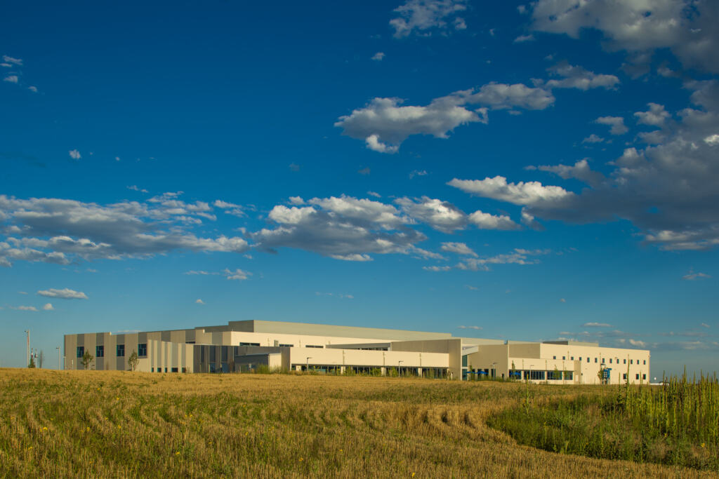 Modern beige industrial building in a wide open field under a vibrant blue sky with scattered clouds, Nordson Value Plastics facility in Colorado.