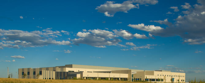 Modern beige industrial building in a wide open field under a vibrant blue sky with scattered clouds, Nordson Value Plastics facility in Colorado.