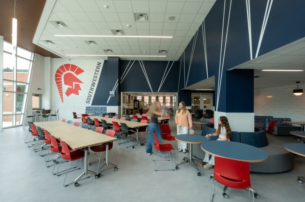 Southwestern Central High School Students socializing in the cafeteria, tall windows providing natural lighting, sets of red chairs at long tables, and a helmeted warrior design on the wall.