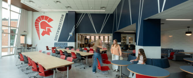 Southwestern Central High School Students socializing in the cafeteria, tall windows providing natural lighting, sets of red chairs at long tables, and a helmeted warrior design on the wall.