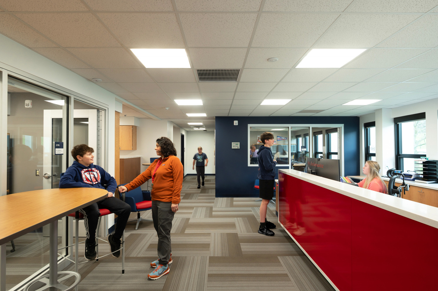 In a brightly lit office at Southwestern Central High School, various students walk, socialize, and talk with a woman behind a striking red reception desk. This was part of Southwestern Central School District Capital Improvements.