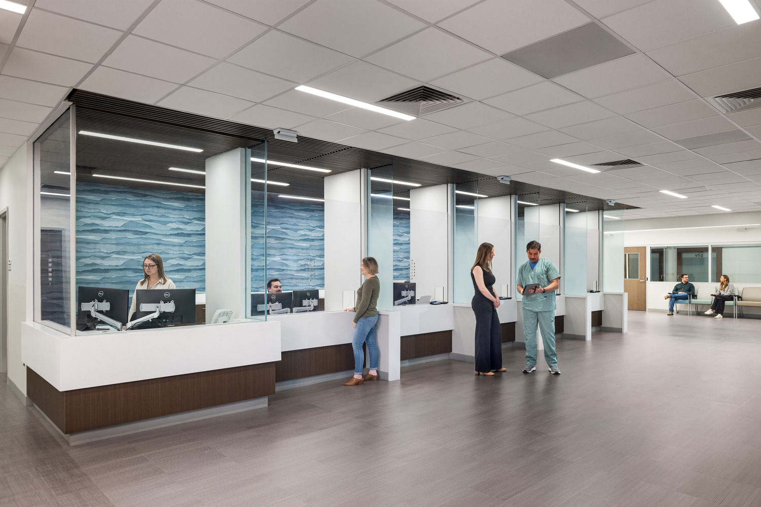 A reception desk with a calming wave pattern behind several employees at monitors, and a few people waiting and talking in the spacious room.