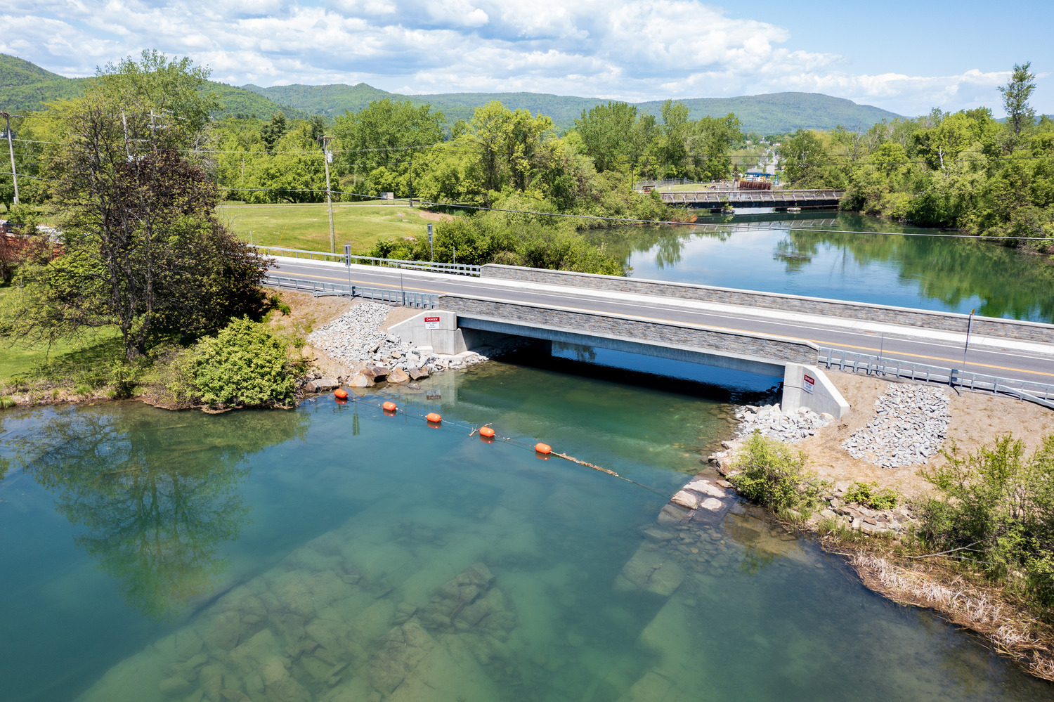 An overhead view of a two-lane bridge crossing a beautiful blue river, and the Adirondack mountains in the distance.