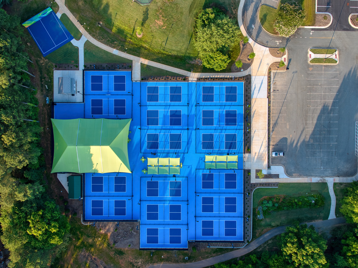 The blue and green pickleball courts from directly above, surrounded by trees on one side and a parking lot on another.
