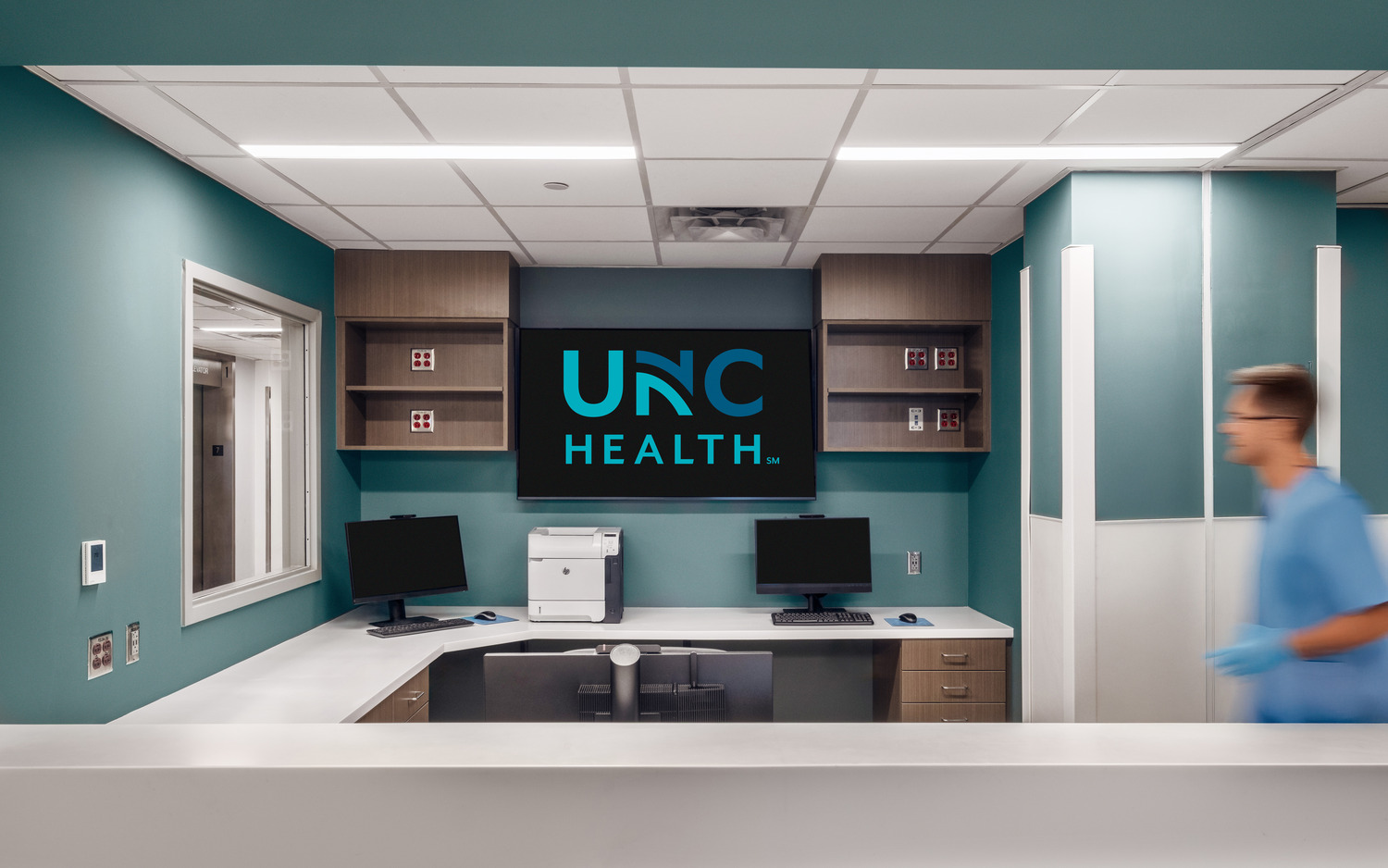 Wayne UNC Healthcare – 7th Floor Reno A monitor with the UNC Health logo at the head of a reception area. A medical worker in scrubs heads towards a small window looking out the side wall of the space.