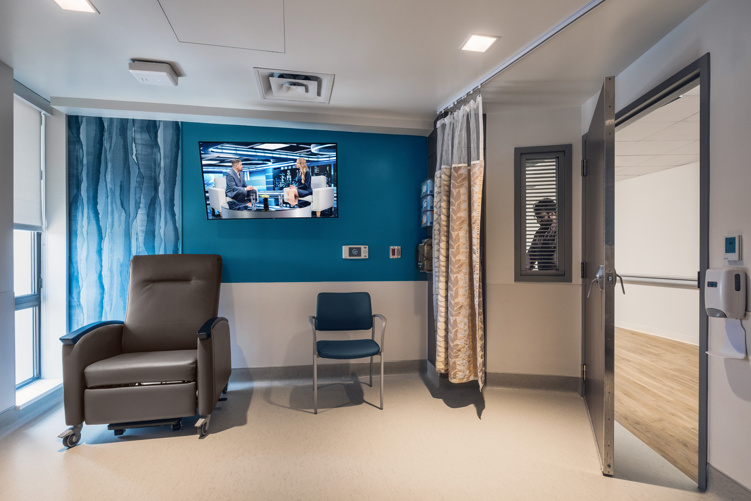 Guest seating and a television against a calming turquoise wall in a Wayne UNC Healthcare patient room.