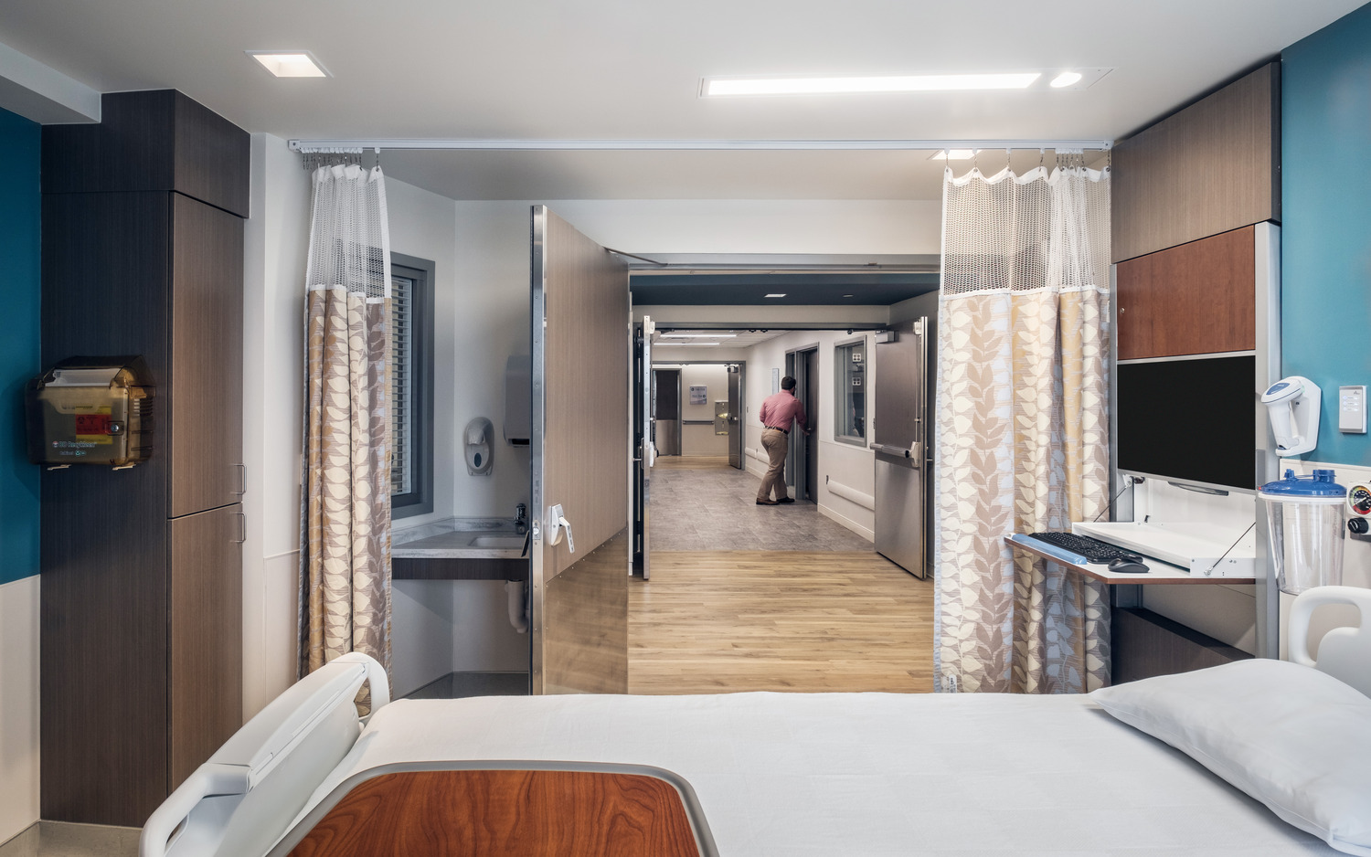 Open doors into a patient room with a medical bed at Wayne UNC Healthcare. A man in a pink shirt and khakis opens a door down the hall.