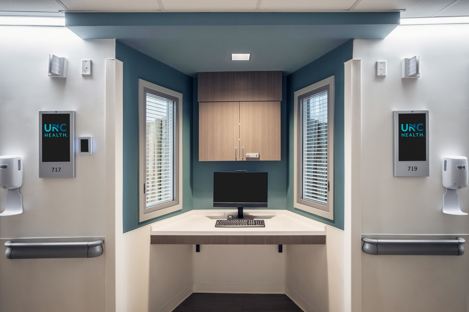 A decentralized nurse station with a computer monitor and shuttered window view into two patient rooms.