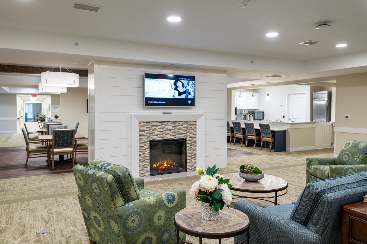 Village_of_St_Edward_at_Wadsworth_Lounge Spacious, cozy lounge with patterned armchairs, a blue couch, and a modern fireplace. Wall-mounted TV above; open kitchen and dining area in the background.