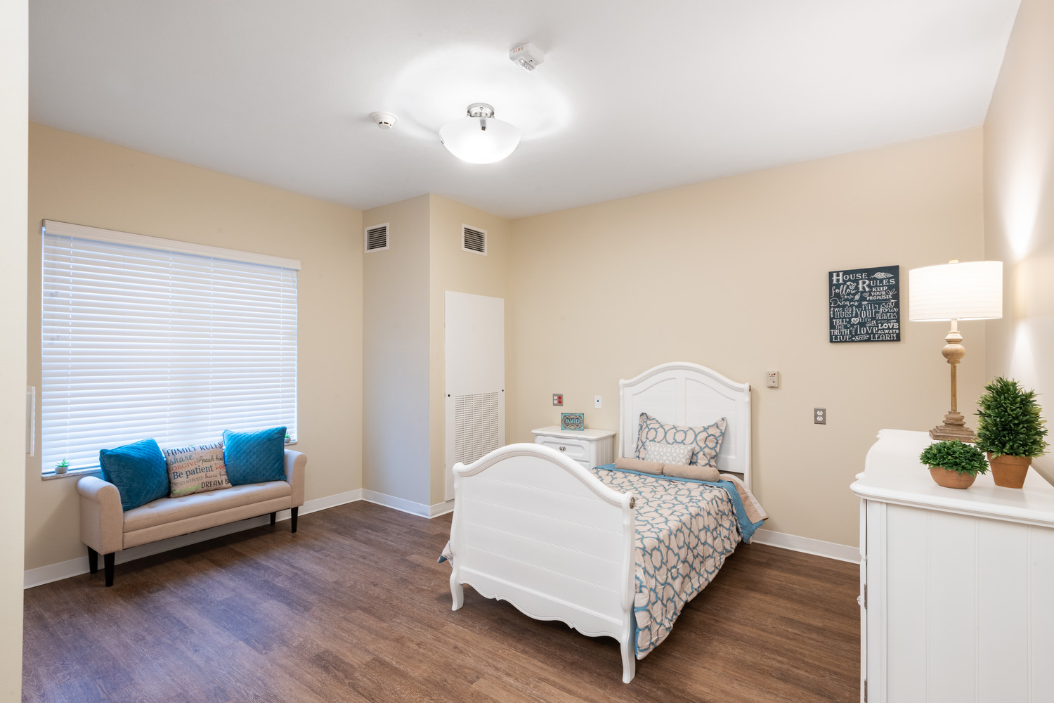 AND_2019_011_Village_of_St_Edward_N34_medium A cozy bedroom at the Village of St. Edward with beige walls and wooden flooring. It features a single white bed with patterned bedding, a small couch with blue pillows, and a dresser with plants.