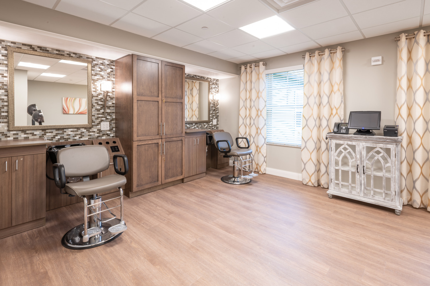 AND_2019_011_Village_of_St_Edward_N36_medium Modern salon room at The Village of St. Edward with two styling chairs facing mirrors and wooden cabinets. Light wood flooring, patterned curtains, warm lighting, and relaxed ambiance.