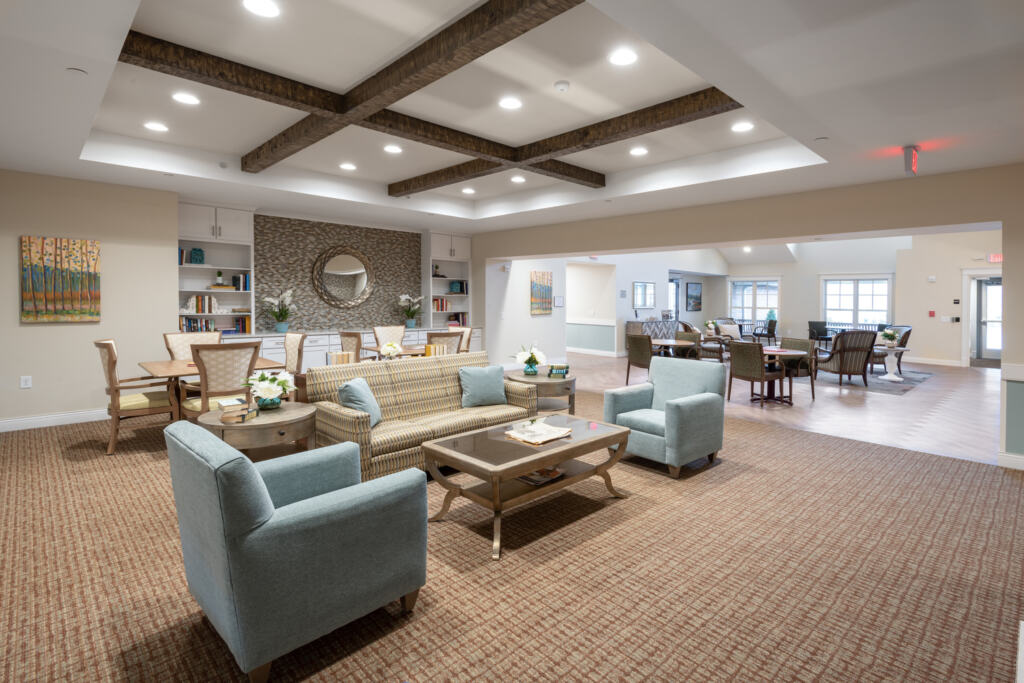 Spacious lounge area with soft lighting, featuring blue armchairs, a beige sofa, wooden ceiling beams, a long dining table, and bookshelves. Cozy and inviting atmosphere.