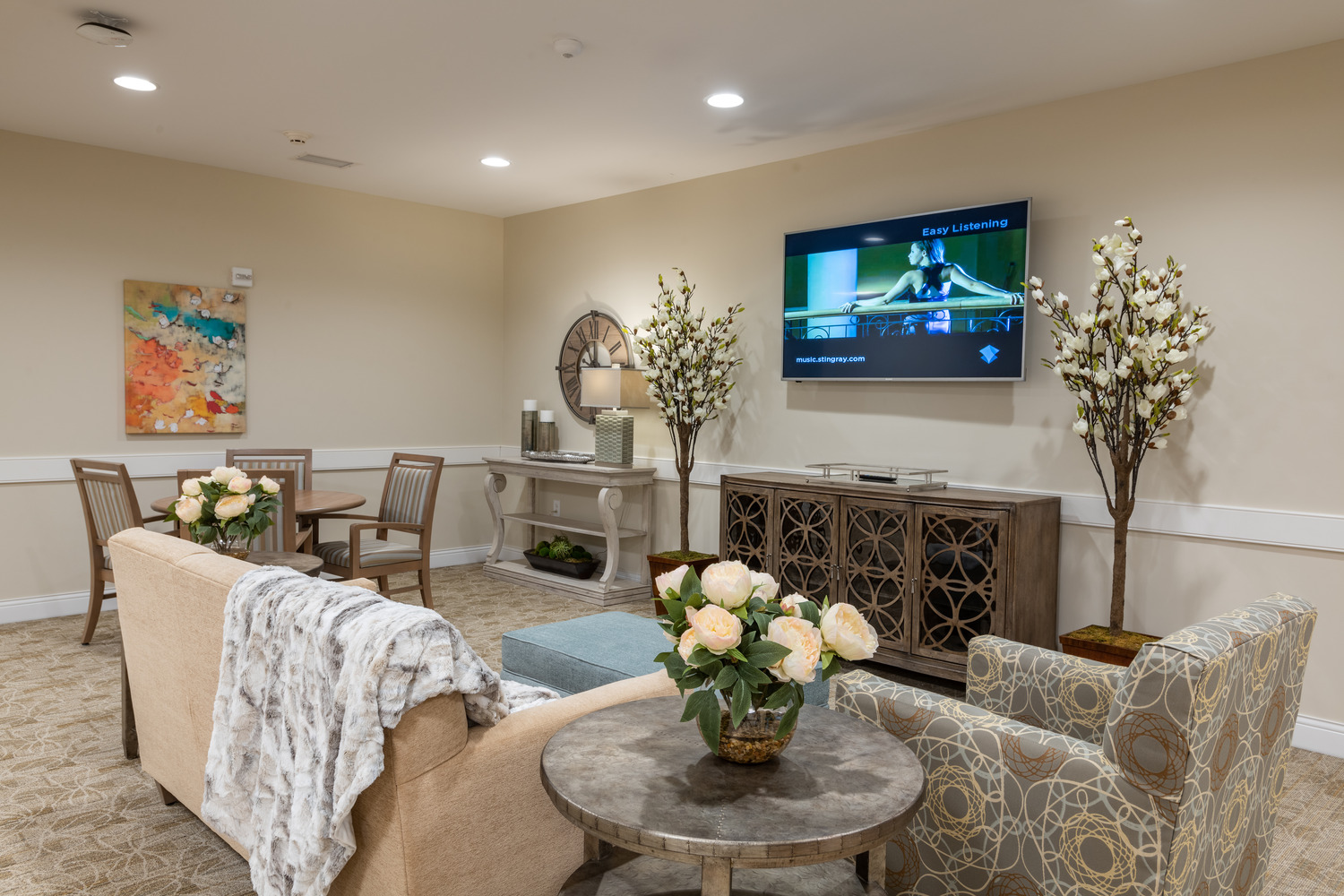 AND_2019_011_Village_of_St_Edward_N46_medium Village of St. Edward at Wadsworth interior showing a cozy living room with beige walls features a sofa draped with a blanket, floral cushions, a patterned armchair, and a TV displaying a show. A console table with lamps and floral arrangements adds elegance.