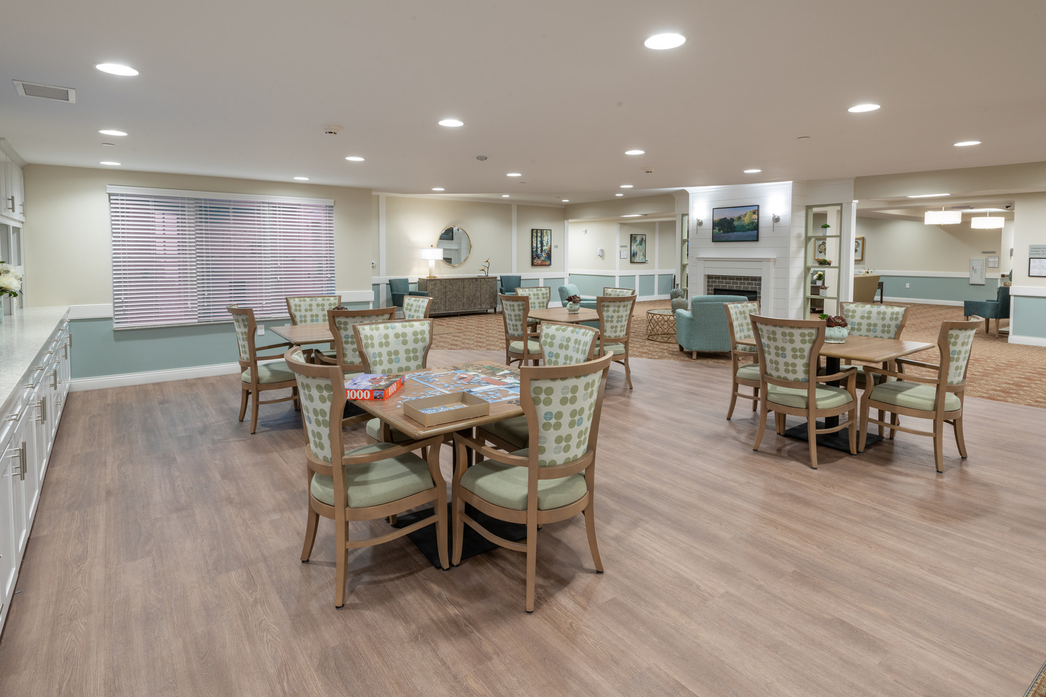 AND_2019_011_Village_of_St_Edward_N48_medium Spacious and well-lit communal room with several wooden tables and green cushioned chairs. A puzzle is on a table, and sofas are near a fireplace.