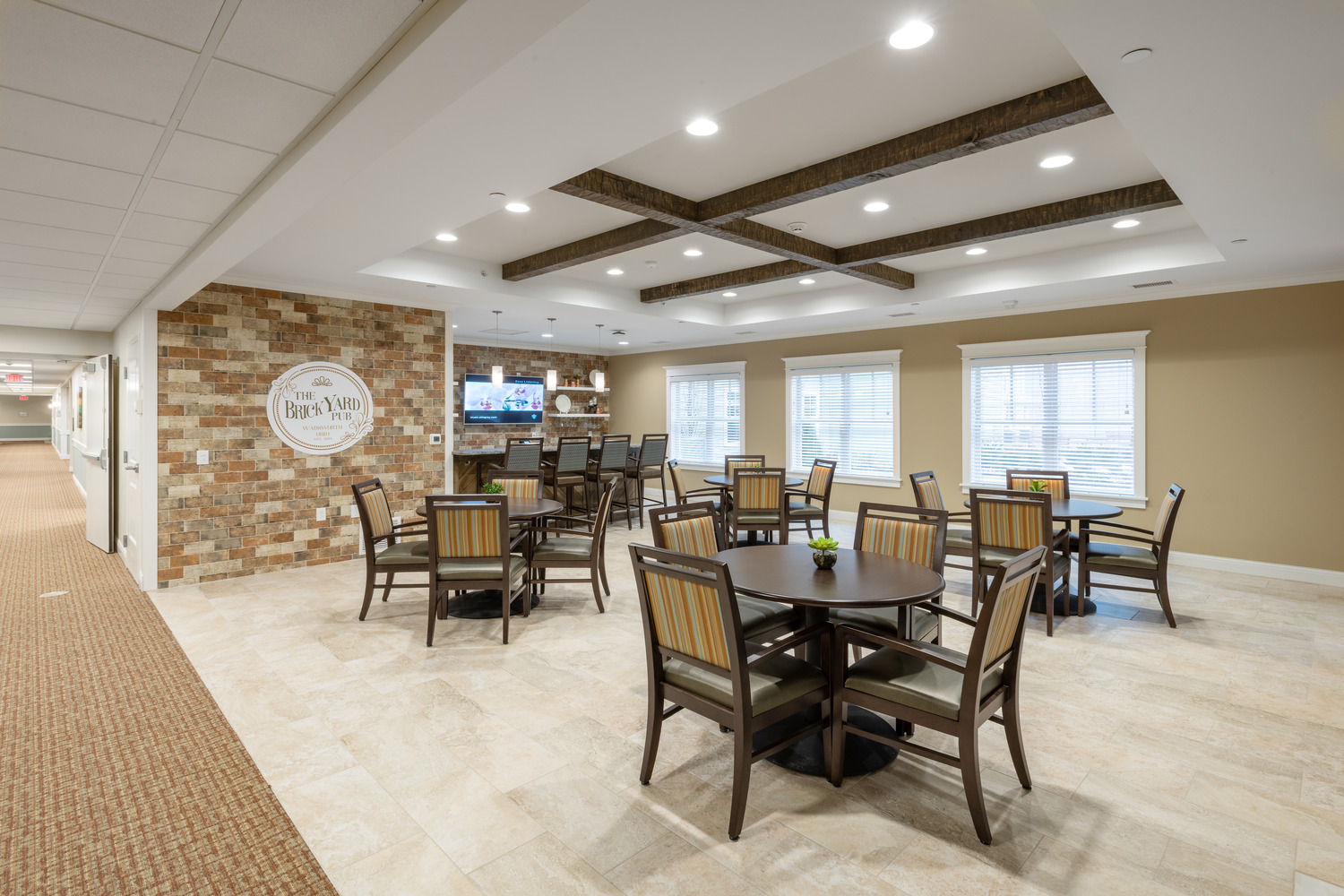 AND_2019_011_Village_of_St_Edward_N6_medium A dining area at the Village of St. Edward at Wadsworth showing a bright dining area with round wooden tables, cushioned chairs, and a brick accent wall, under a ceiling with exposed beams. Cozy and inviting atmosphere.