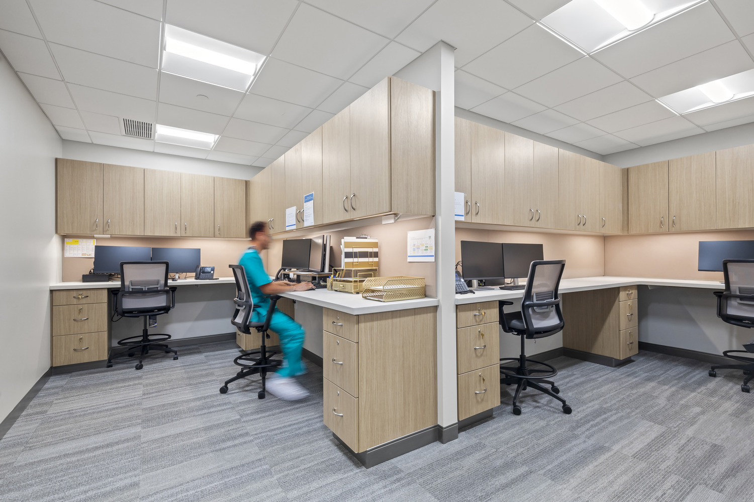 Backlit counters with sets of monitors and overhead cabinets, a person in scrubs sitting at one of the computers.