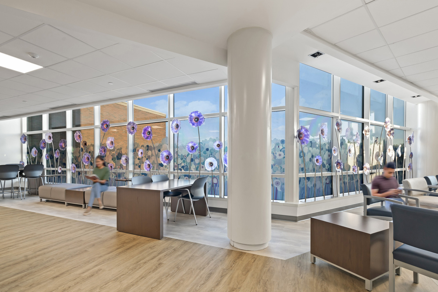 A few people sit and read in Westerville Cancer Center's waiting area. Chairs line a wall of windows decorated with purple glass flower designs.