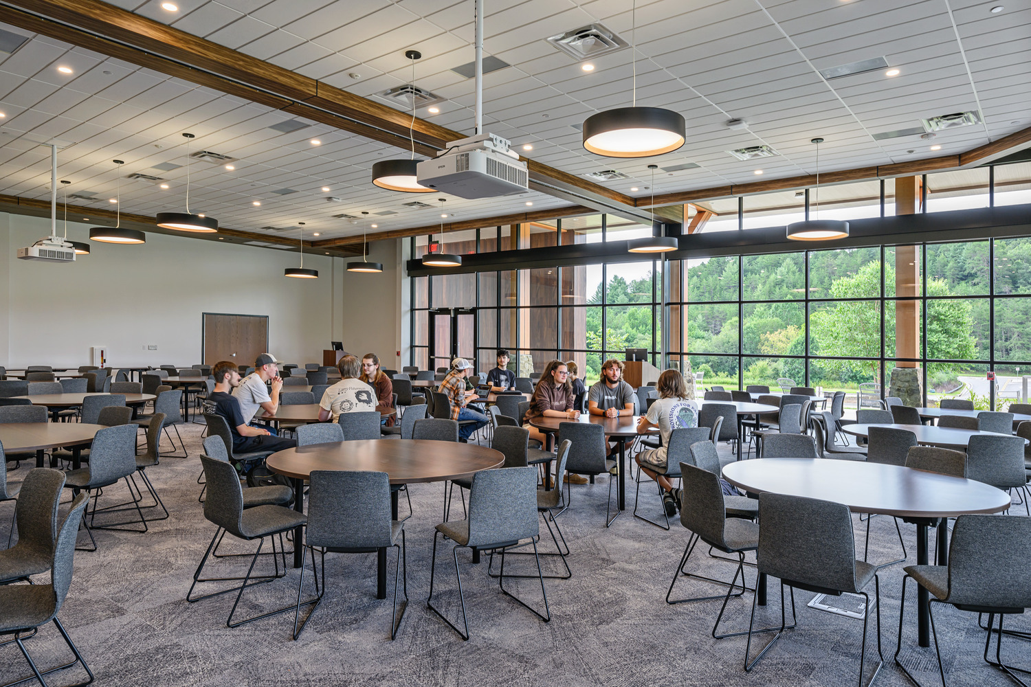 A spacious University dining hall with circle tables and rows of windows on the far wall, students sitting and talking.