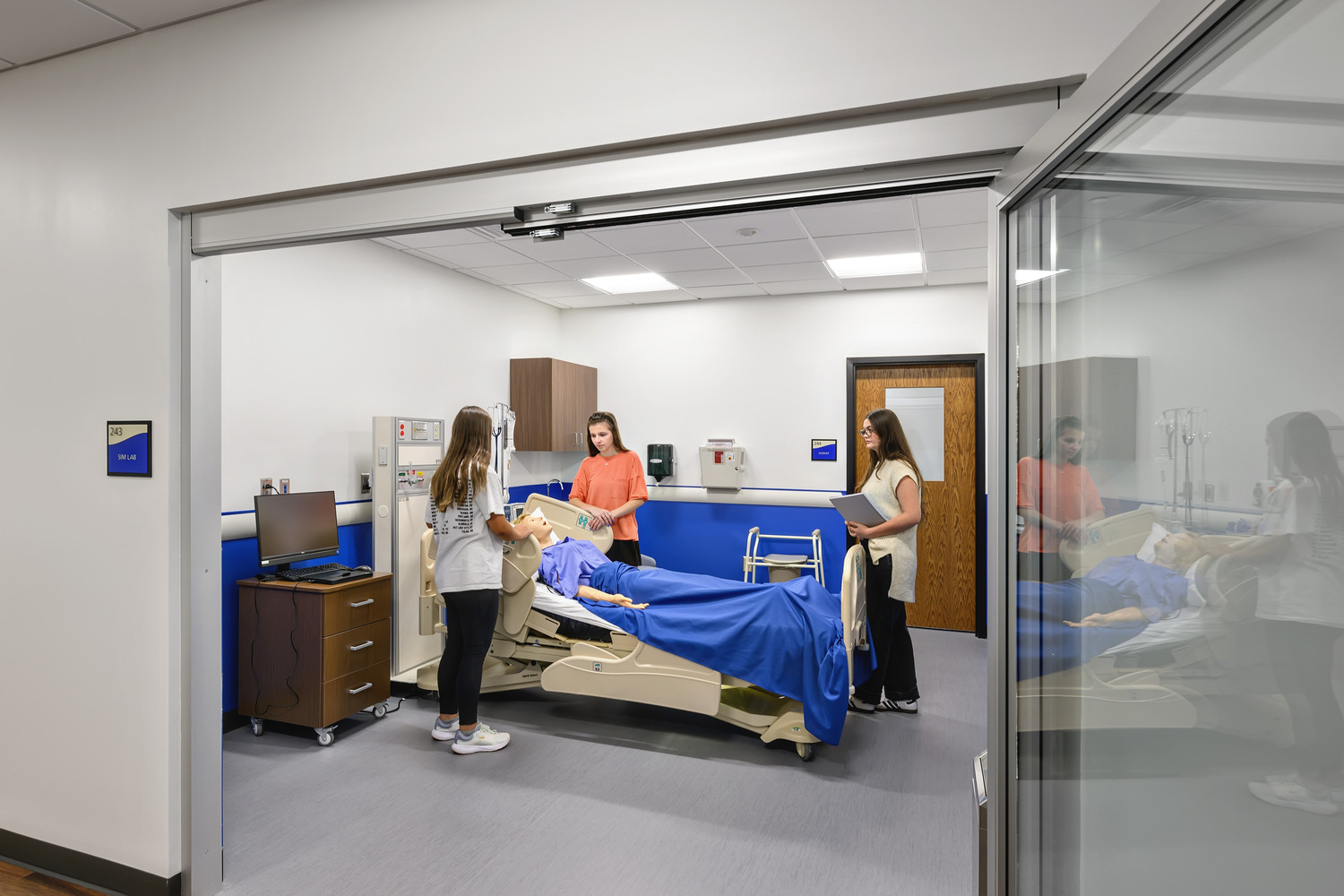 An open glass door looking into a medical education suite, students surrounding a medical bed with a human patient simulator.