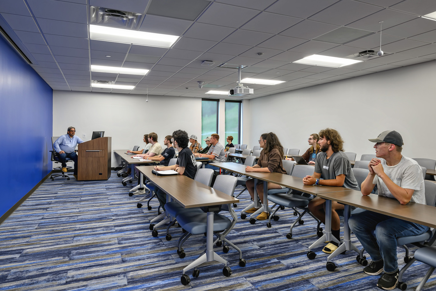 A classroom with rows of tables and students attentively listening to a professor at the head of the room.