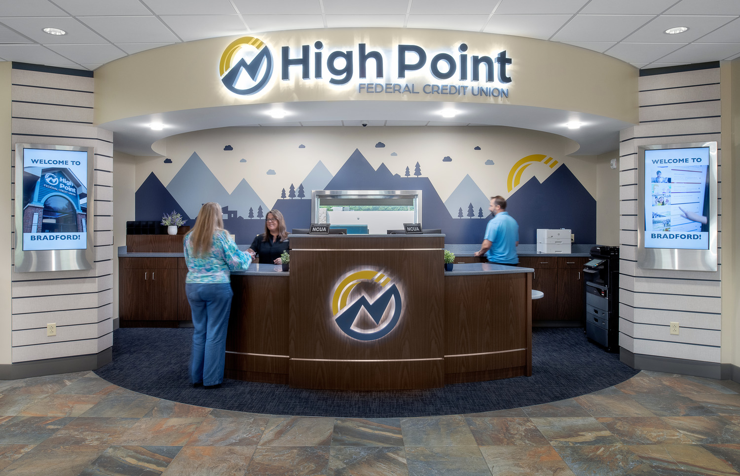 A rounded reception desk with High Point Federal Credit Union on the bulkhead above, the HPFCU logo on the desk, and soft blue mountain designs on the back wall.