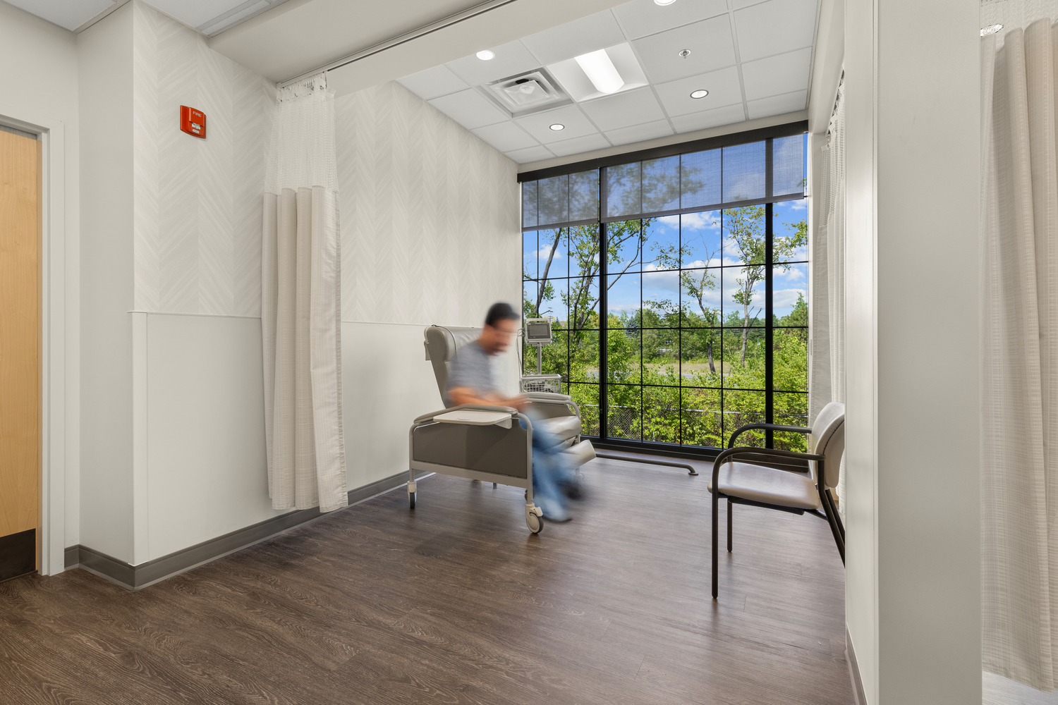 A patient of Vive Vascular sits in a chair in front of a floor-to-ceiling window in a medical bay with an open curtain.