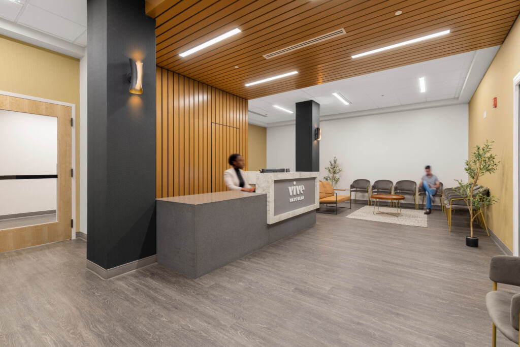 The front desk and waiting area at Vive Vascular where a man waits and reads a book and a woman types on a keyboard at reception.