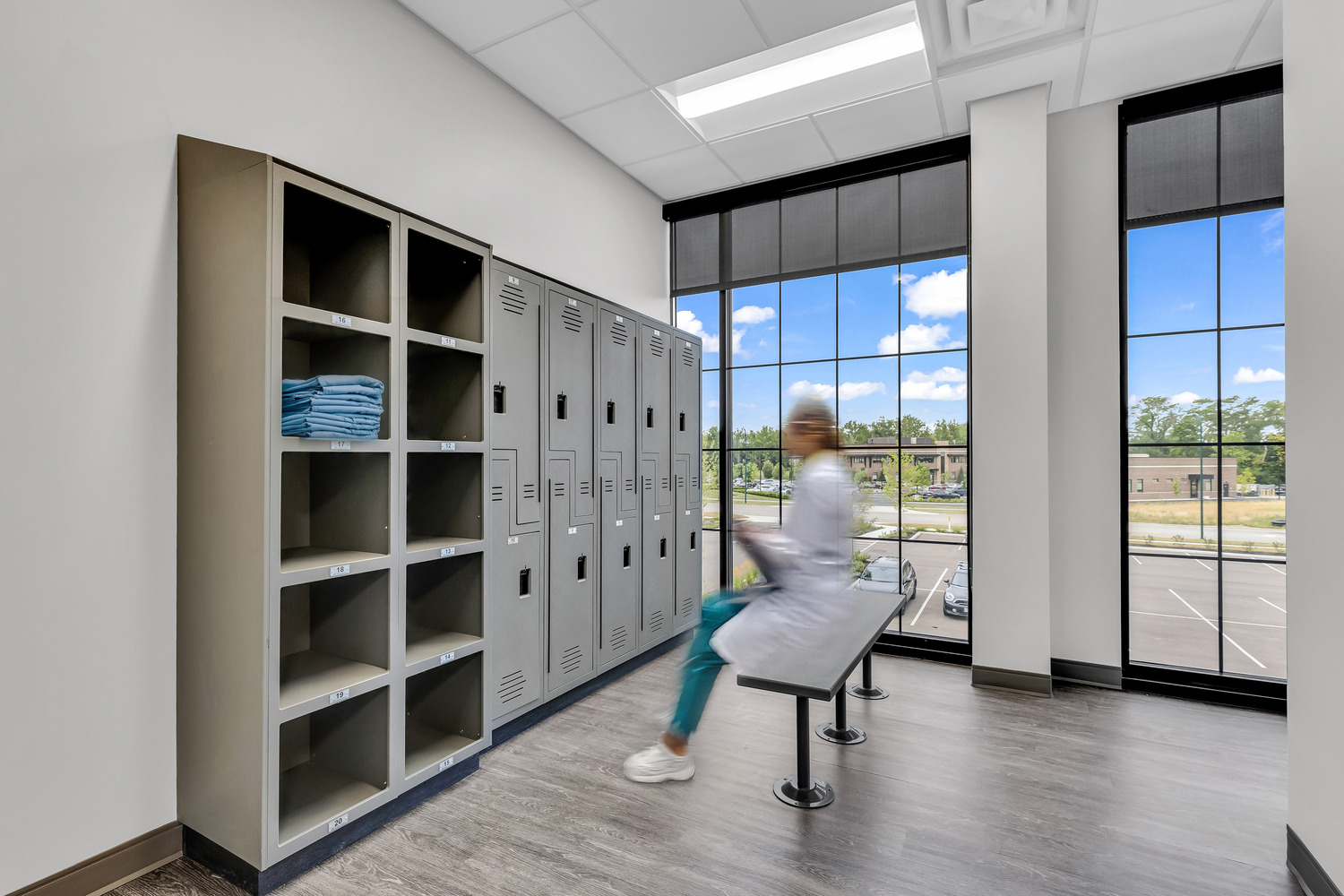 A medical professional sits on a bench in front of a set of item lockers next to tall windows.