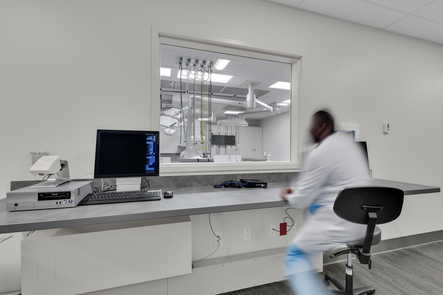 A doctor sits at a counter, looking through a square window to a surgical suite.