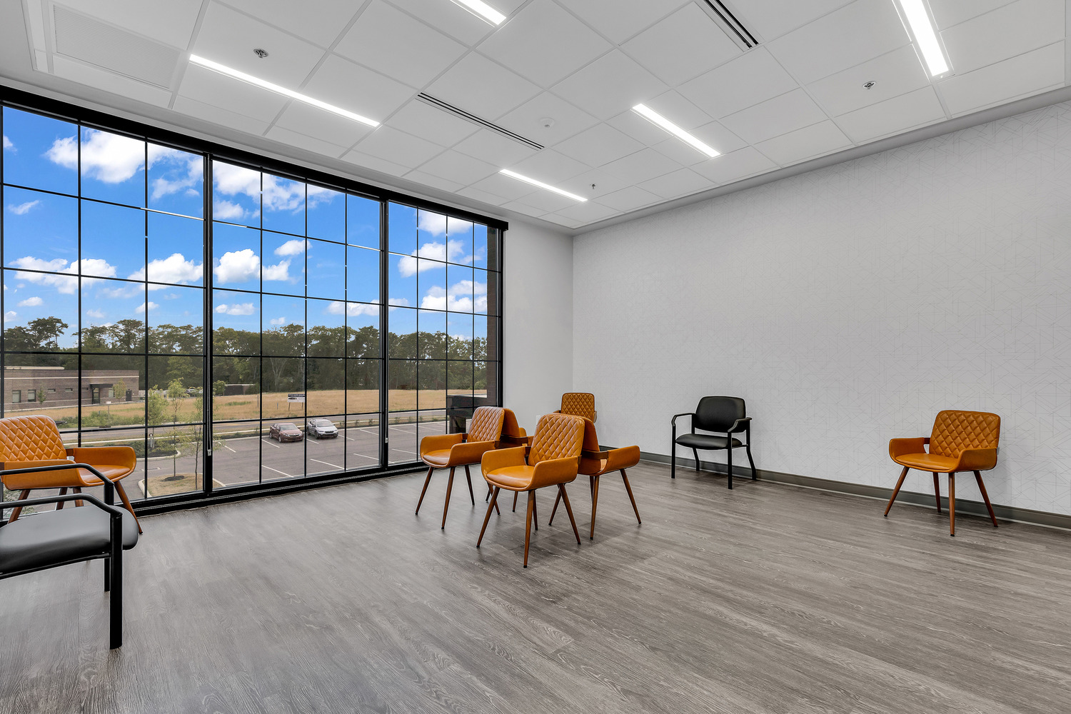 A waiting area with various chairs and tall windows overlooking the parking lot under a sunny blue sky at Trillium Surgery Ambulatory Surgical Center (ASC).