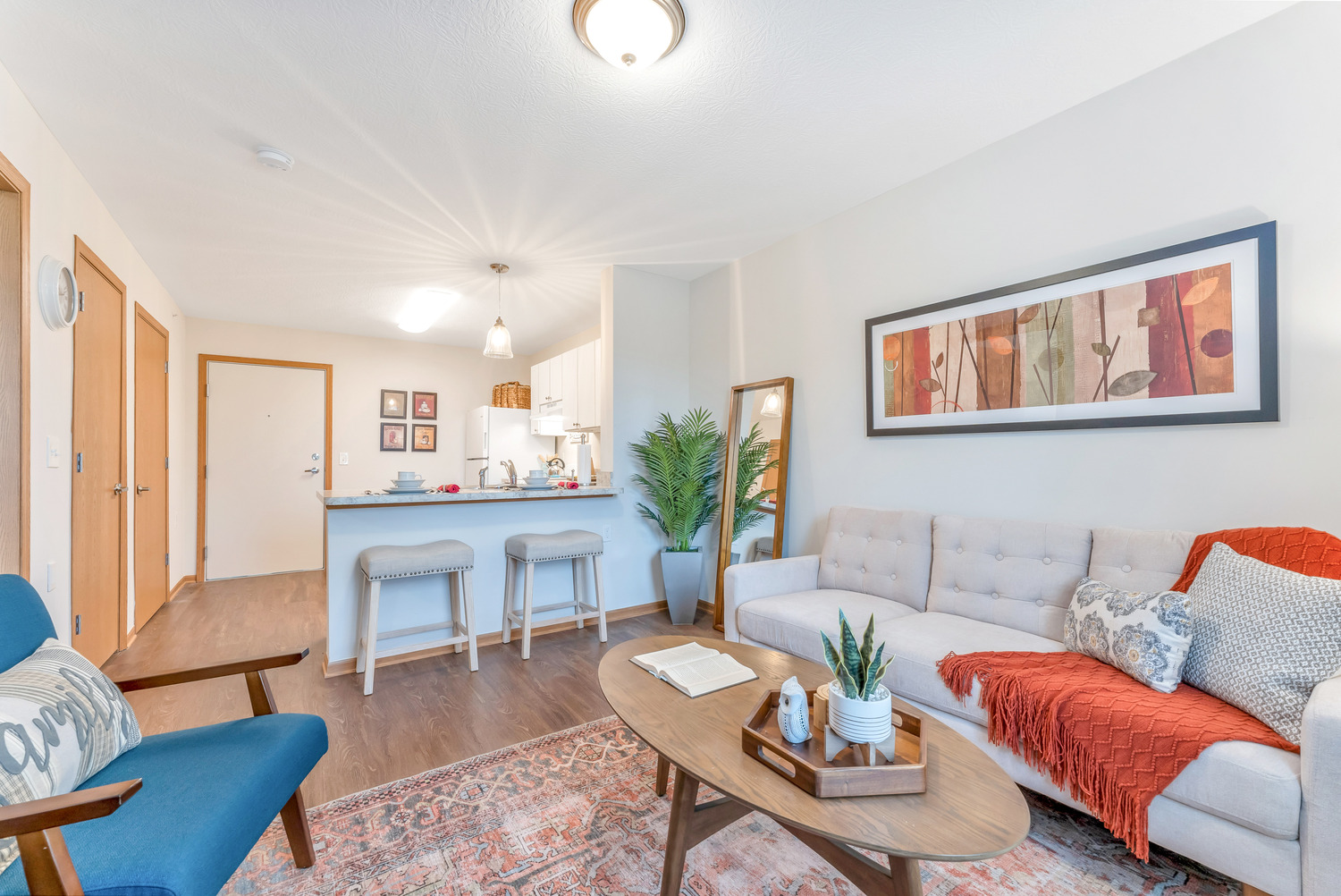 In one of Homestead Village Active Senior Living Facility's residential units, a kitchen with a bar and stools connects to a living room with colorful, comfortable furniture and a coffee table.