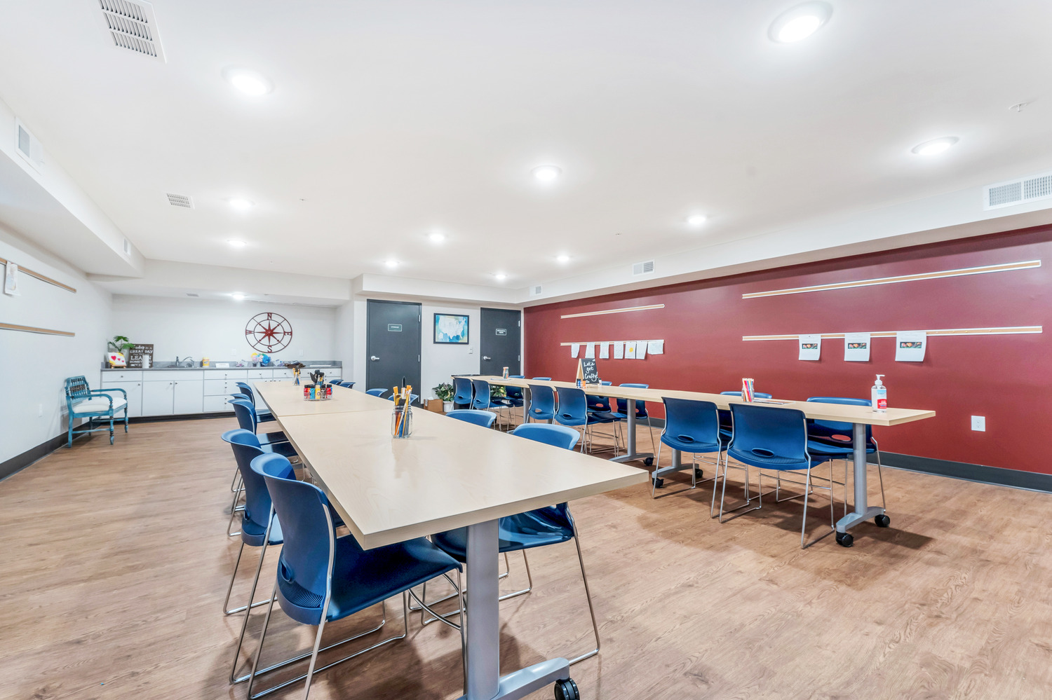 An activity room with a red accent wall, long tables with rows of chairs and cups of paint brushes and art supplies inside Homestead Village Active Senior Living Facility.