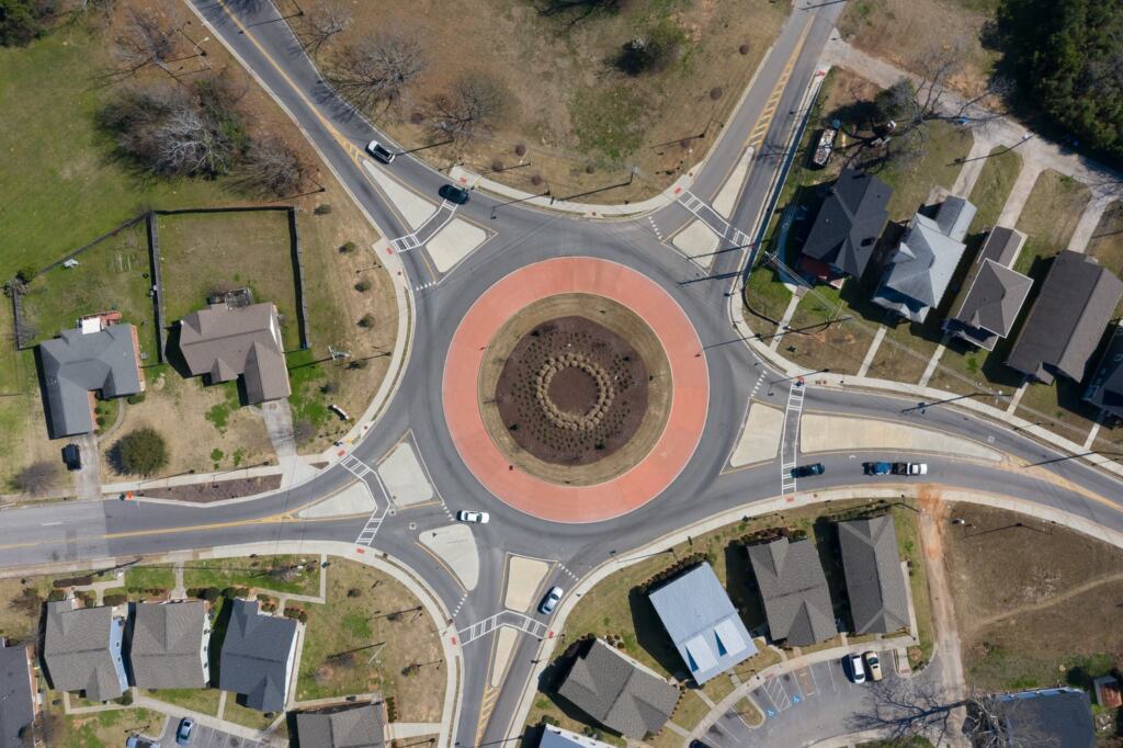 Aerial view of Twiggs Rd at Wrightsboro road showing a suburban roundabout with a red circular center, surrounded by houses and lawns. Cars navigate the intersection, suggesting calm suburb life.