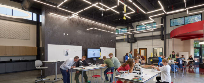 This presentation space in a Middle/High School STEAM wing is designed to facilitate interactive project showcases. The classroom is open concept with dark gray walls, red accent colors, wall-mounted TVs and movable chairs and tables. College-Ready Campuses: Hospitality-Inspired K-12 School Design