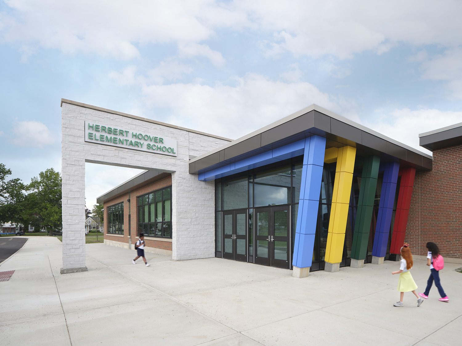 Modern school entrance with colorful columns and glass doors under a blue sky. Children walk along the path, creating a lively, cheerful atmosphere.
