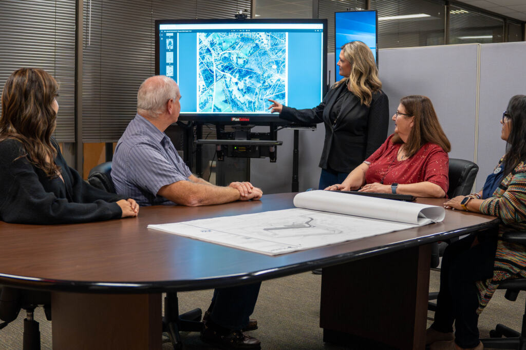 a woman is pointing to a map while standing in front of people seated at a table as part of CPL's on-call government services.