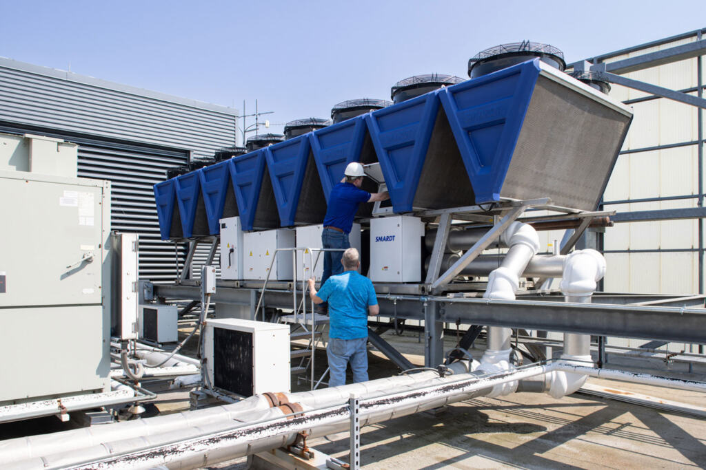 Two CPL technicians work on large industrial HVAC units with blue covers on a rooftop. One person is on a ladder while the other assists from the ground.