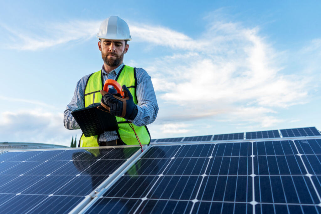 Technician inspecting rooftop solar panels with a clamp meter and tablet, representing how energy incentives support the implementation and maintenance of renewable energy systems.